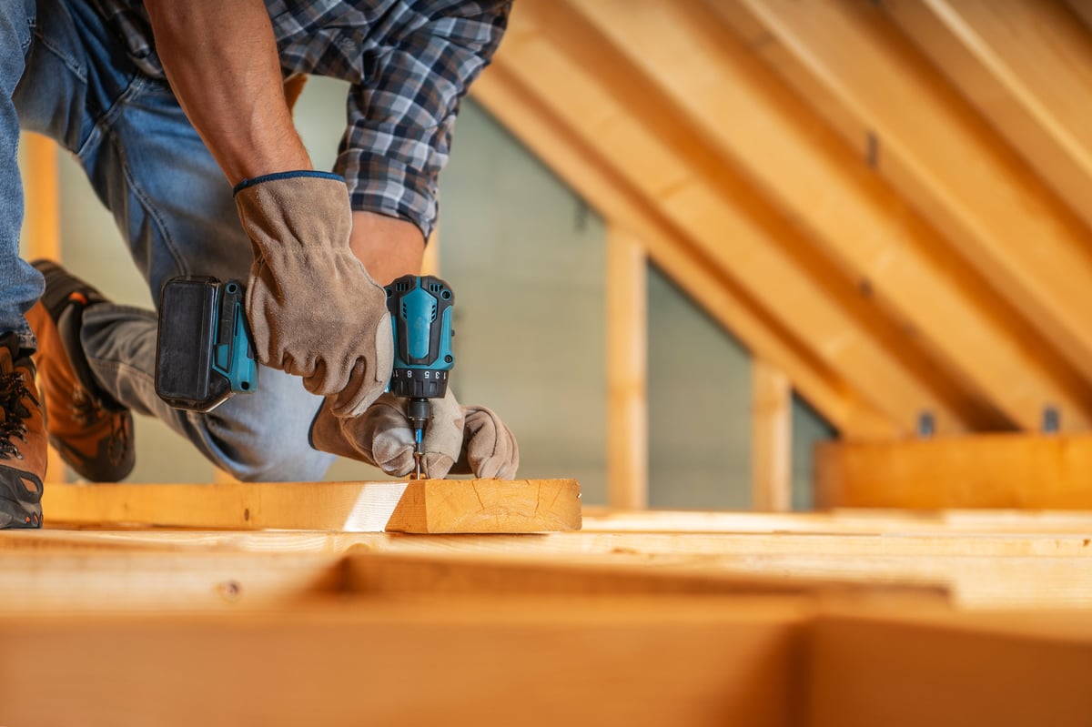 A skilled carpenter works diligently in an attic, securing wooden beams with a power drill while wearing gloves
