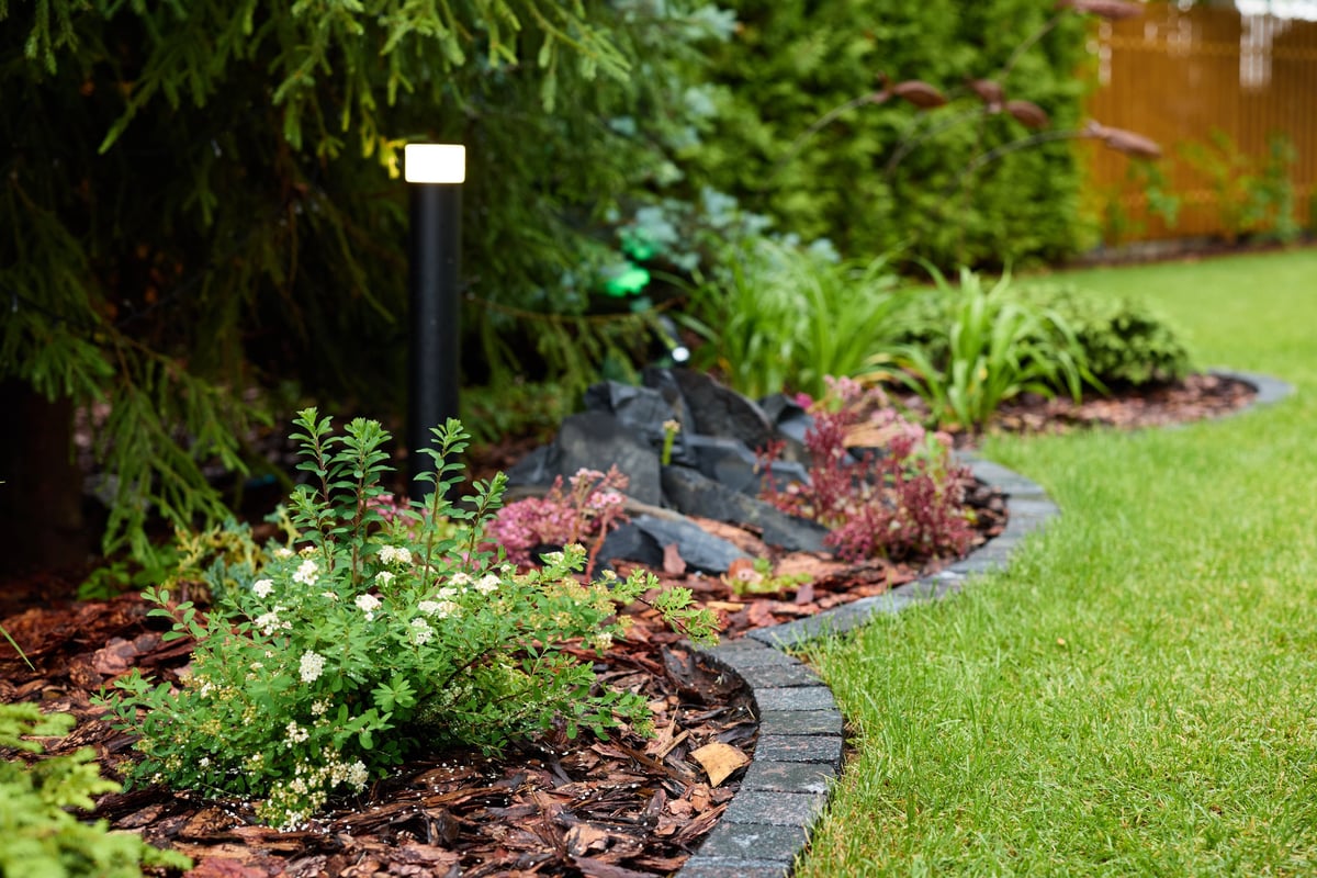 Lush landscaped garden bed with a curved stone border, decorative rocks, flowering plants, and a manicured lawn