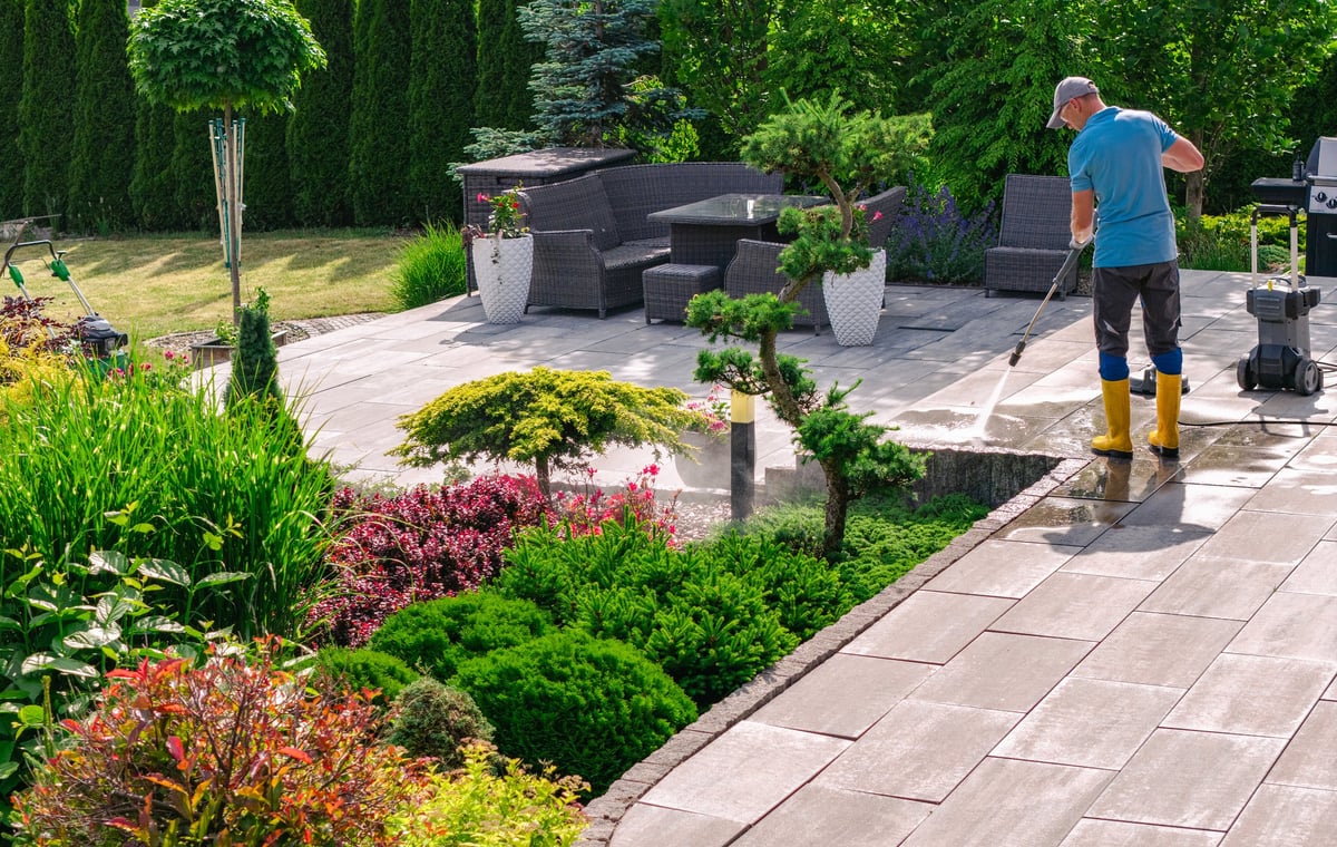 Gardener pressure washing a paver patio surrounded by lush plants, decorative shrubs, and wicker outdoor furniture in bright sunlight