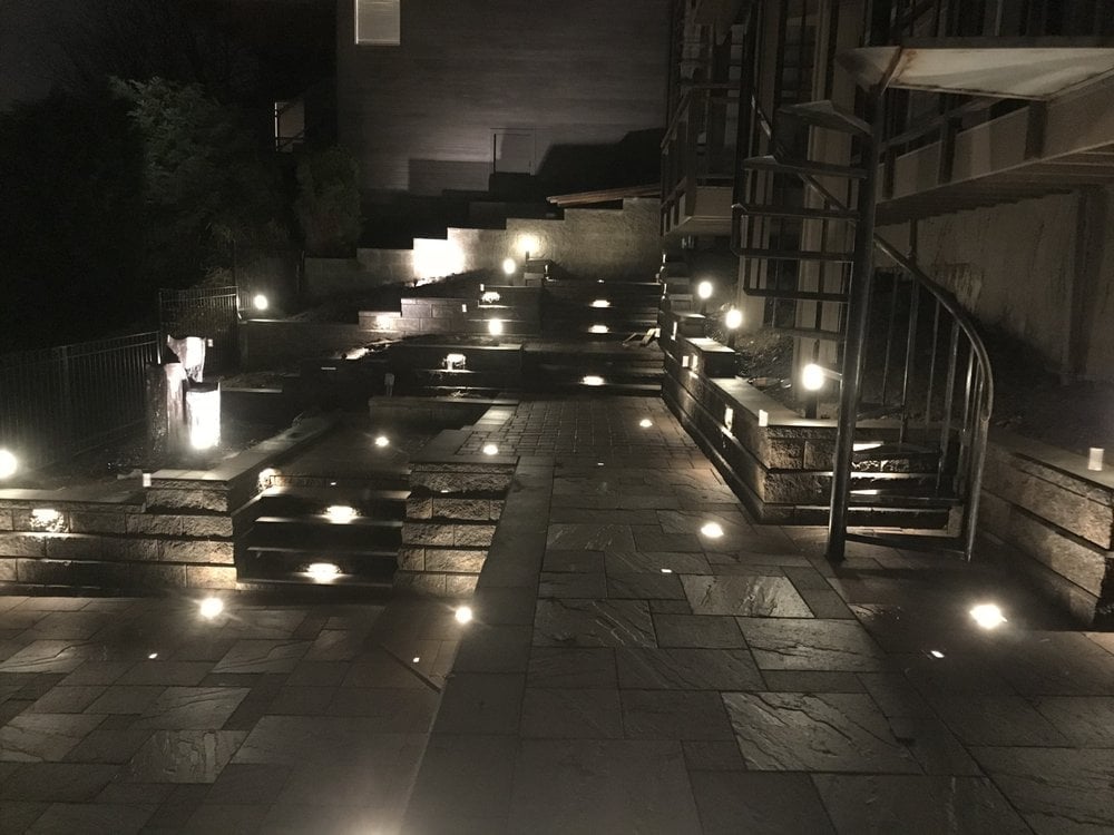 Night view of a modern courtyard paved with stone, lit by recessed floor lights and warm wall-mounted fixtures