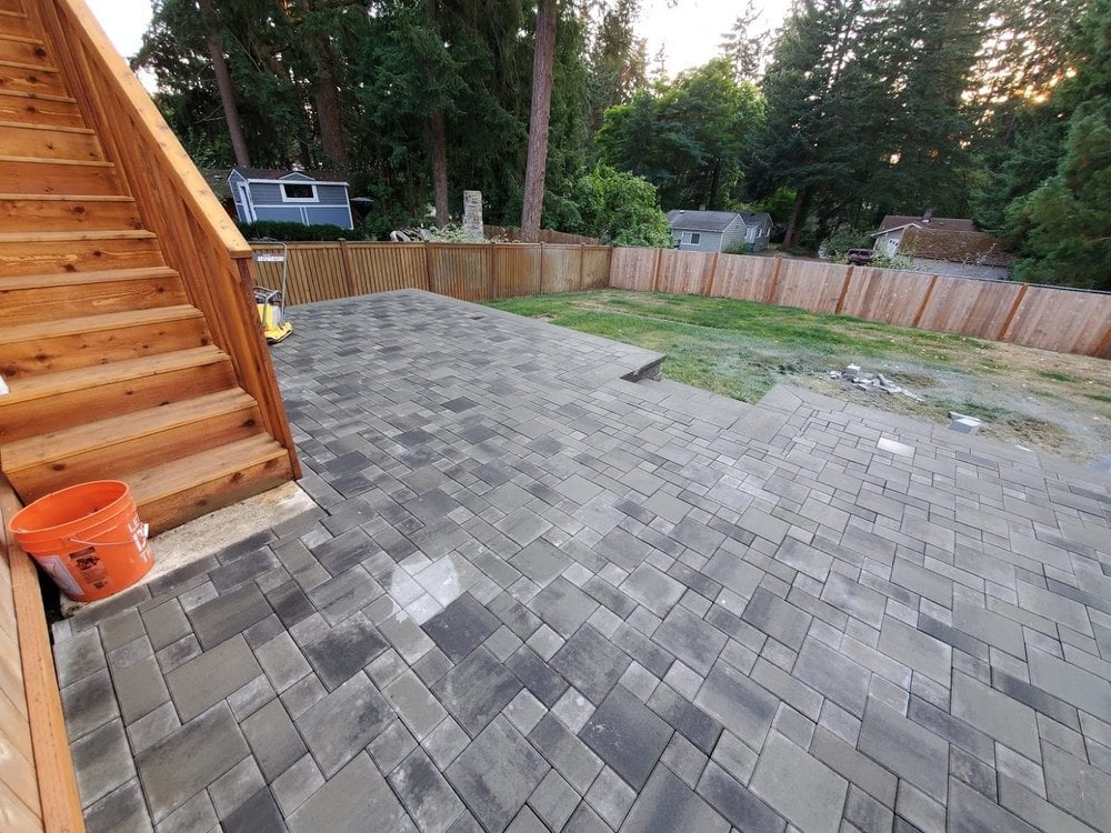 Backyard patio with gray stone pavers next to a wooden deck, wooden fence, and tall trees