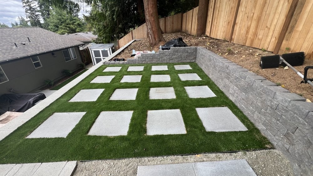Backyard patio with concrete pavers on artificial turf, bordered by a block retaining wall and wooden fence