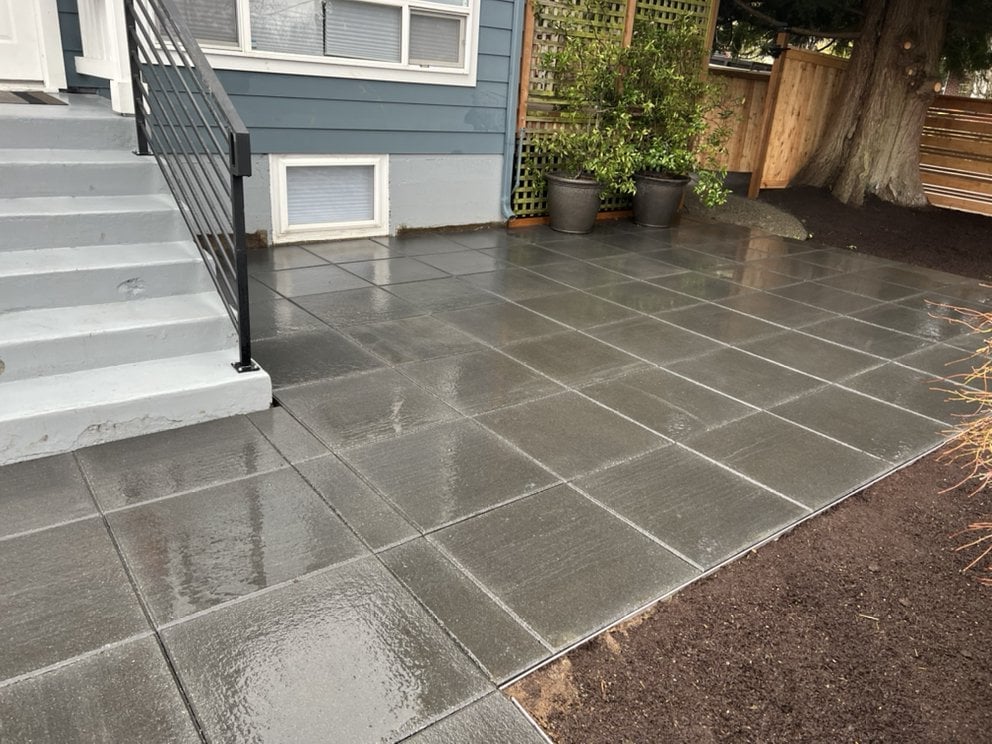 Modern front patio with dark gray pavers, metal railing, potted plants, and wooden fence