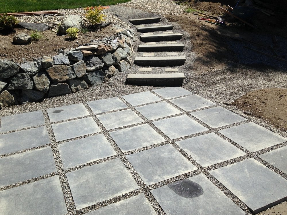 Outdoor patio with large square stone pavers and gravel joints leading to wooden steps and a stone retaining wall