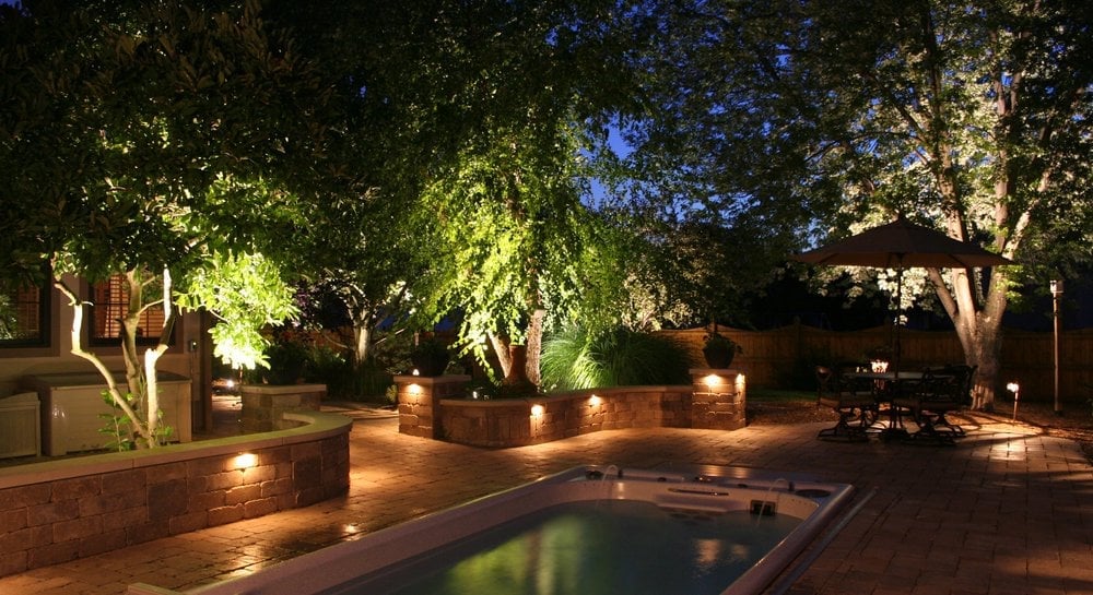 Illuminated backyard at night with a swimming pool and warm landscape lighting among the trees