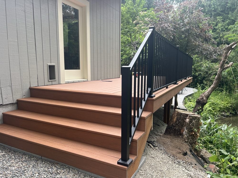 Wooden deck with brown steps leading to a gray house with black railings, surrounded by green trees