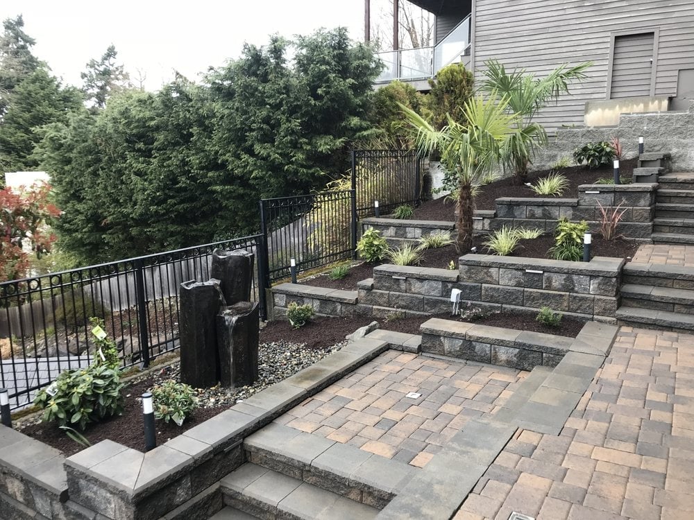 Residential patio with tiered stone planter beds, deck railing, tropical plants, and paver surface