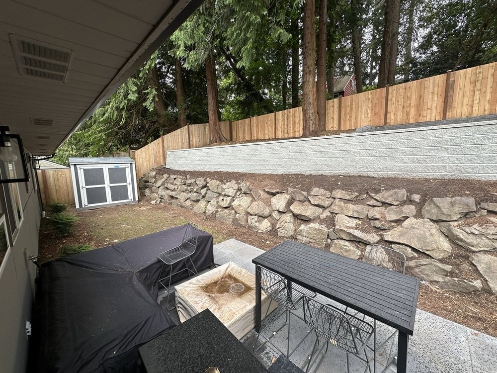 Backyard patio area with covered seating space, stone firepit, wooden fence, and storage structures surrounded by tall trees
