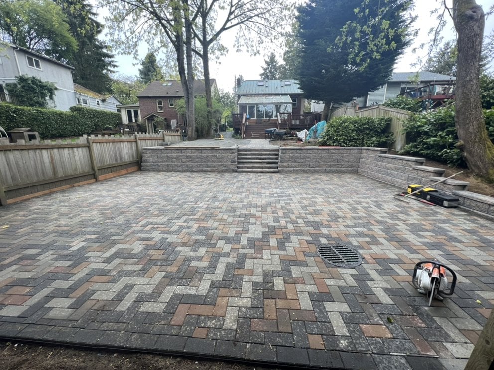 Large paver patio with herringbone-style geometric pattern surrounded by trees and residential fencing