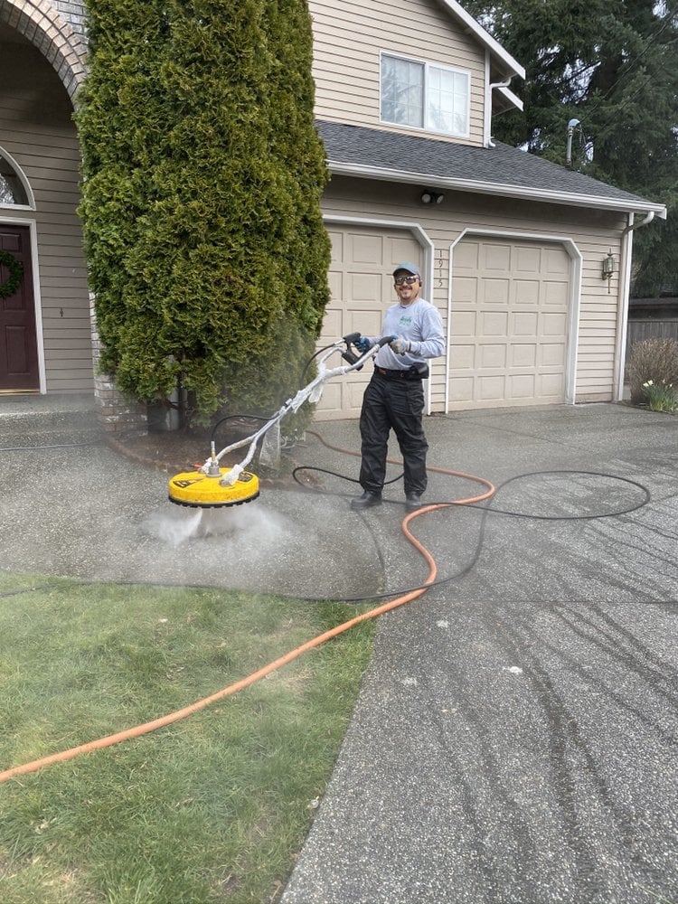 Man pressure washing a driveway with surface cleaner attachment in front of a two-car garage and house