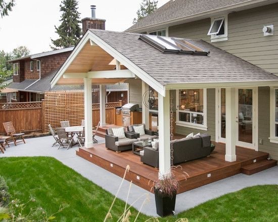 Modern residential patio addition with white columns, wooden deck, seating area, and attached to beige house with green lawn