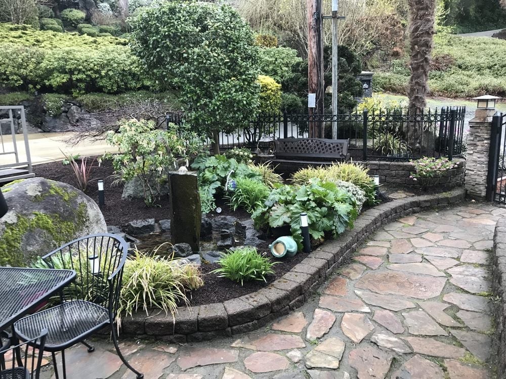 Lush garden patio with stone pathways, curved flower beds with white flowering plants, and black metal chairs