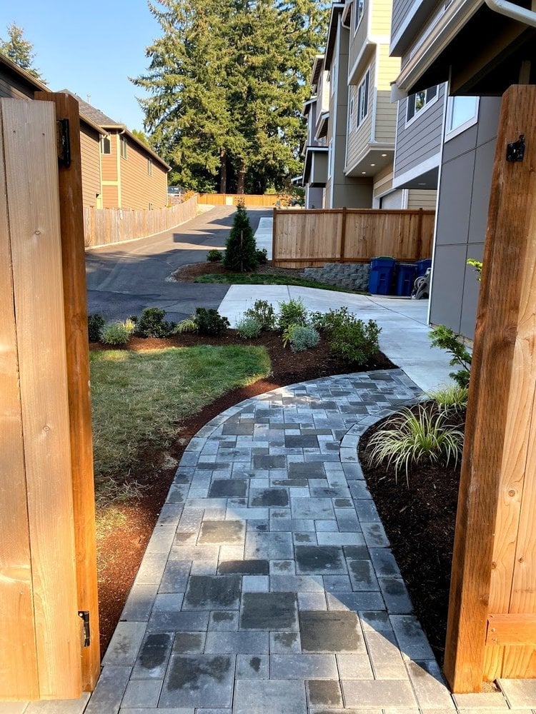Wooden gate opening to a curved paver pathway leading through a residential backyard with landscaping and modern houses